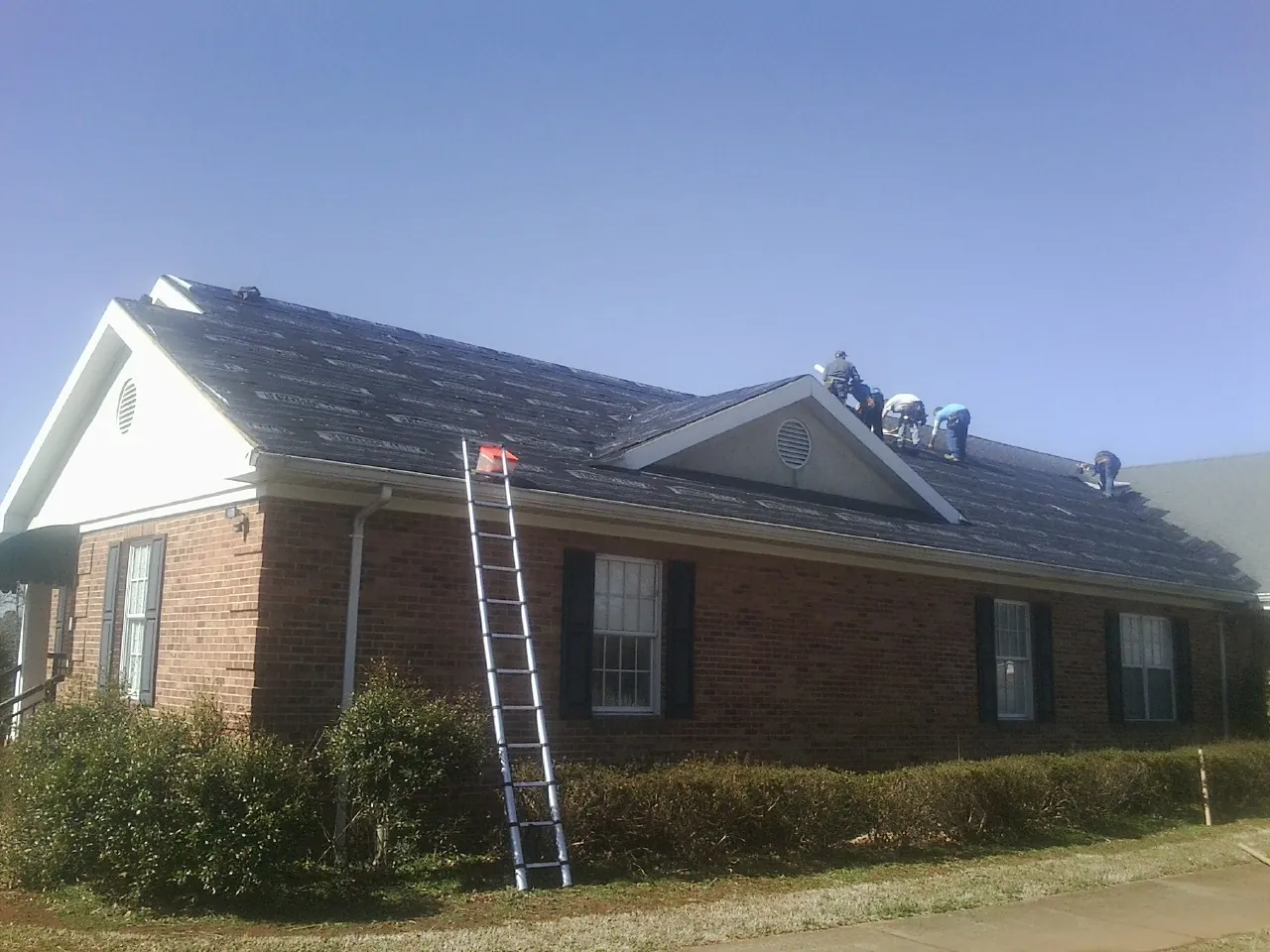 Crew tearing shingles off a brick ranch roof with a ladder set — Metal Roof Repair work by our crew in New Sharon