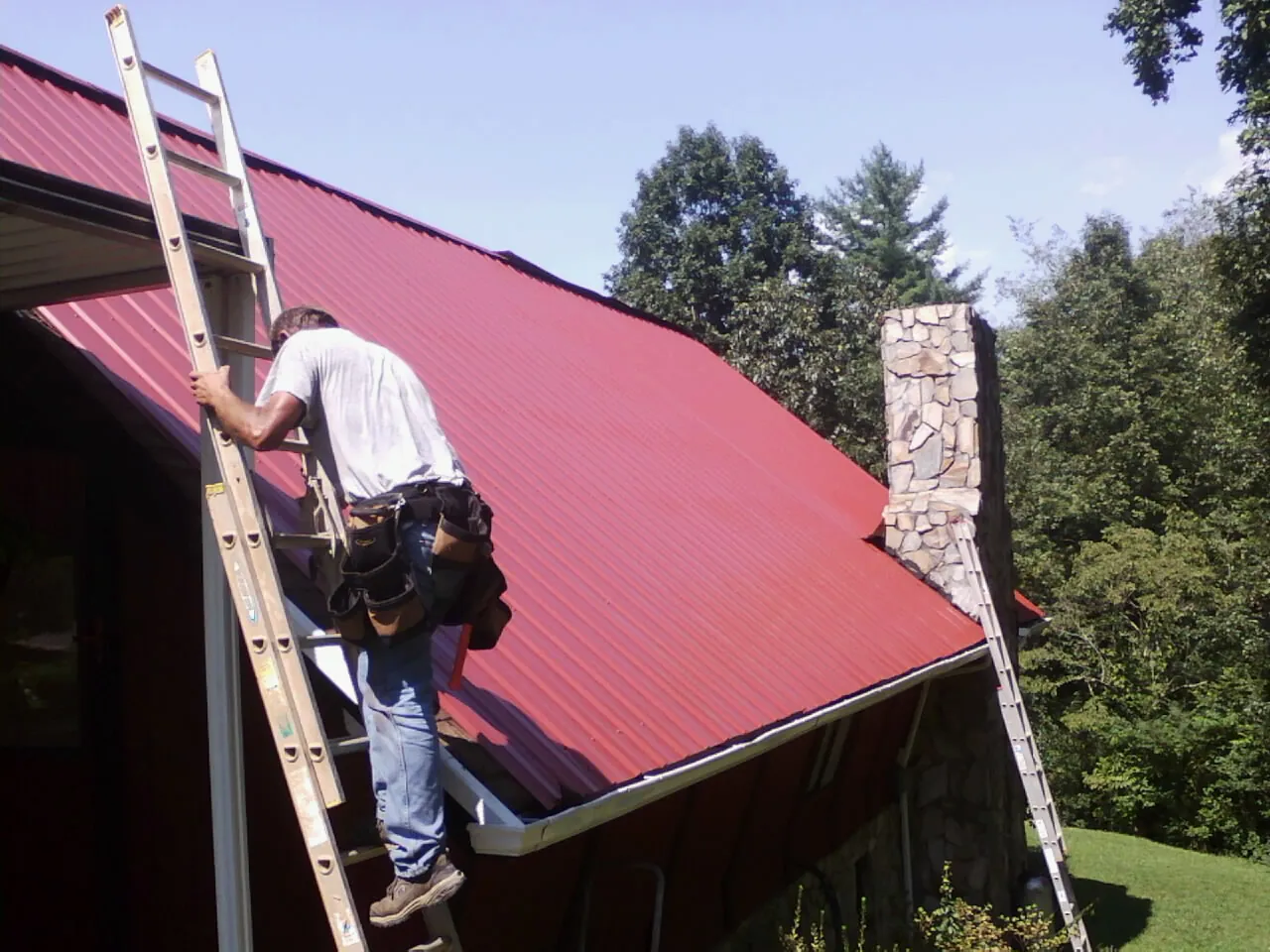 Roofer on a ladder beside a red metal roof and stone chimney — Soffit & Fascia Repair by Roofing Brothers in New Sharon